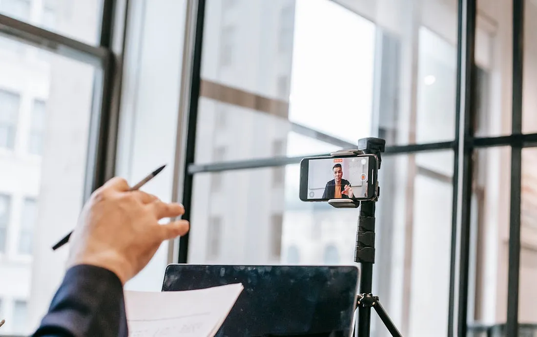 Fotografia de uma pessoa em um ambiente de escritório, participando de uma videoconferência ou gravação usando um smartphone montado em um tripé. A câmera do smartphone está voltada para a pessoa, que aparece na tela do dispositivo enquanto fala ou apresenta algo. A mão de outra pessoa, segurando uma caneta e um pedaço de papel, também aparece em primeiro plano, sugerindo que a pessoa está envolvida na conferência, talvez como espectadora ou participante