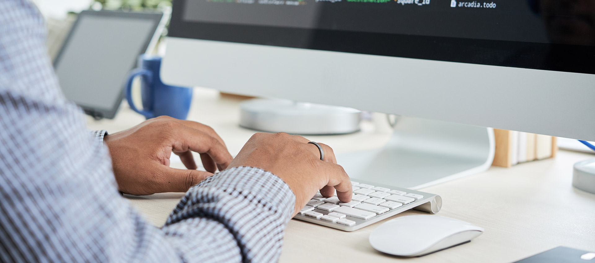 Pessoa digitando em um teclado branco conectado a um computador desktop com uma tela grande. A tela do computador exibe linhas de código. A pessoa usa uma camisa de manga comprida xadrez. No fundo, desfocados, há outros objetos sobre a mesa, como um tablet e uma caneca azul